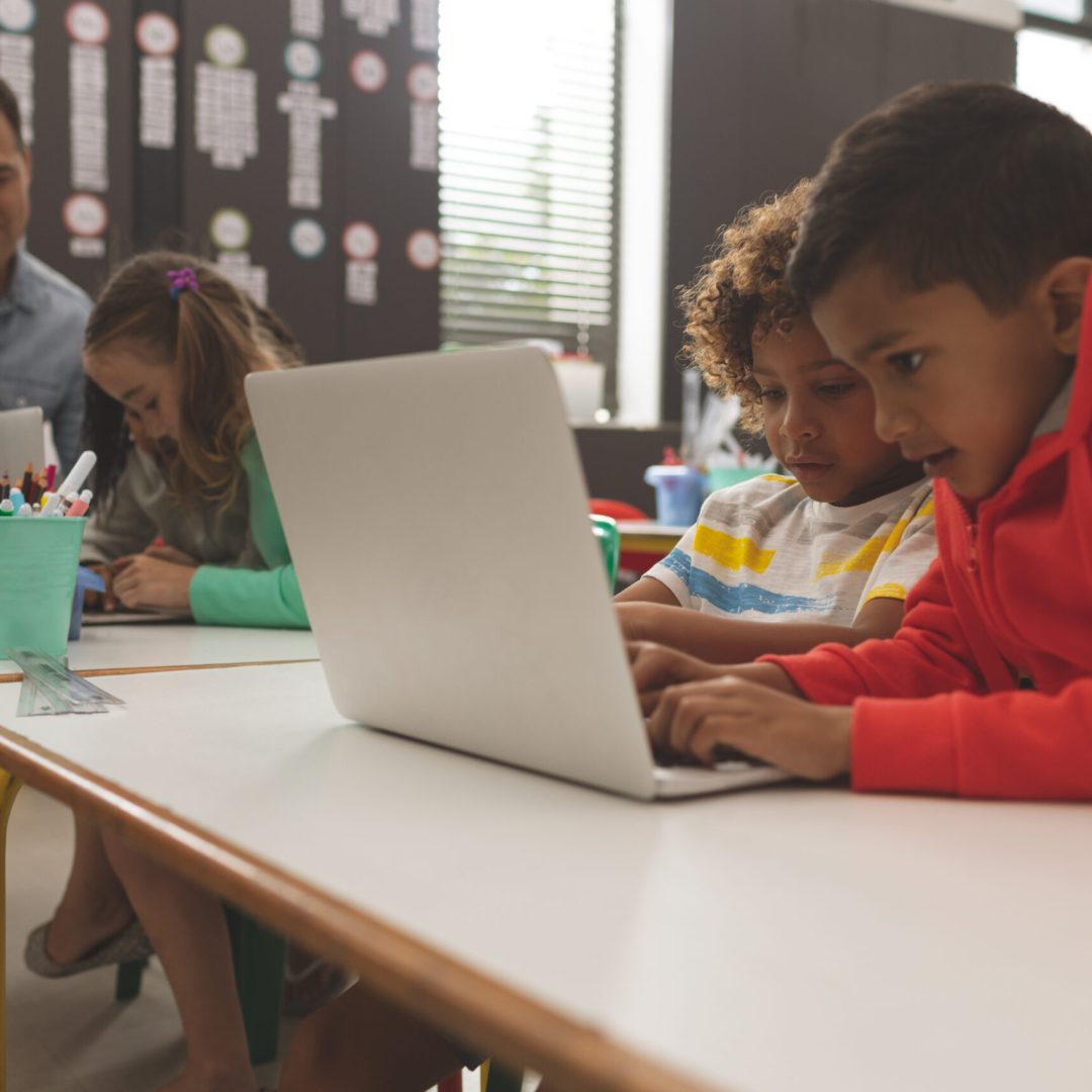 Side view of two mixed-race school boys working and scrolling on one laptop while their teacher teaching to another school girl in background